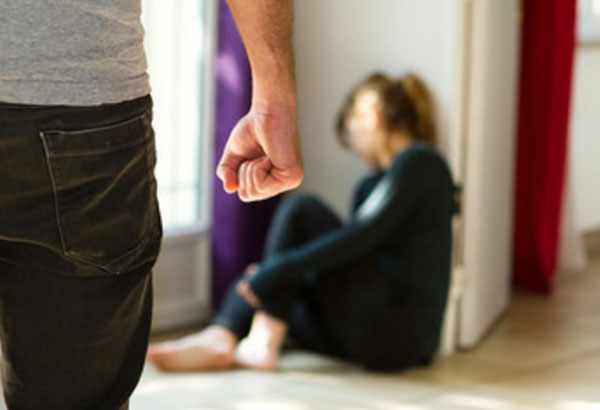 Мужчина с сжатой в кулак рукой приближается к женщине, которая сидит на полу под стеной